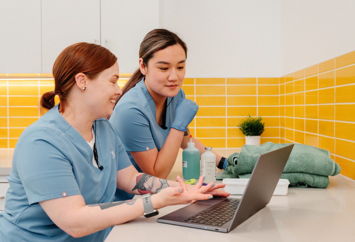 Two technicians looking at a computer. Both are wearing identical light blue scrubs and both have tattoos.