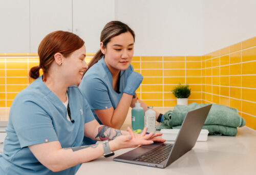 Two technicians looking at a computer. Both are wearing identical light blue scrubs and both have tattoos.